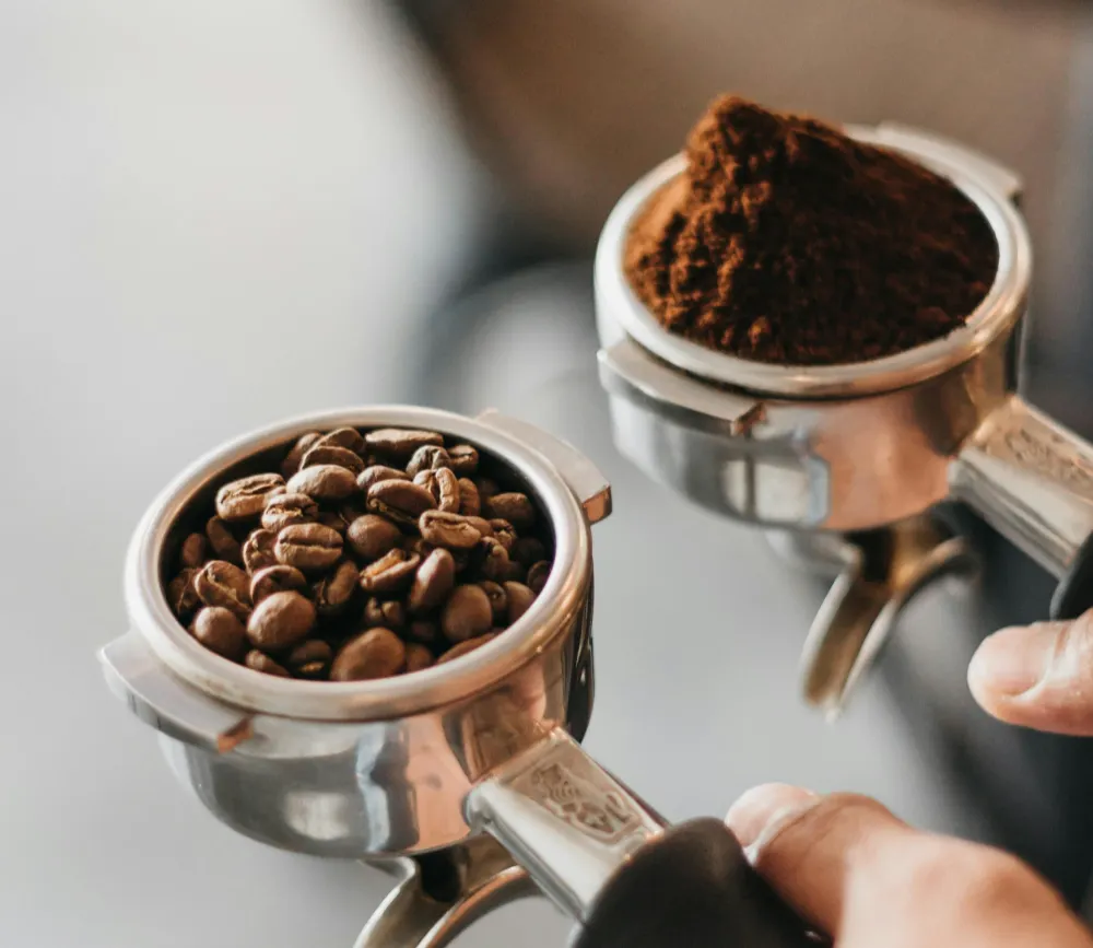  barista prepares espresso using two portafilters—one filled with whole coffee beans, the other with freshly ground coffee—highlighting the coffee preparation process.